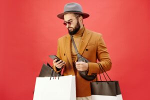 A stylish man wearing a hat and sunglasses checks his phone while holding multiple shopping bags. This image represents how YouPrice helps smart shoppers find premium savings on their purchases through mobile-friendly features and convenient deal alerts.