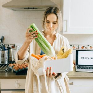 A woman holding fresh vegetables, examining a grocery delivery in her kitchen, showcasing the high-quality, fresh produce delivered by Morrisons right to the customer’s doorstep, ensuring a seamless shopping experience.