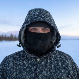 A person wearing a hooded, insulated winter jacket with a face covering, standing in a snowy landscape. Highlights the protective features of Volcano’s jackets, designed to withstand extreme cold and wind.